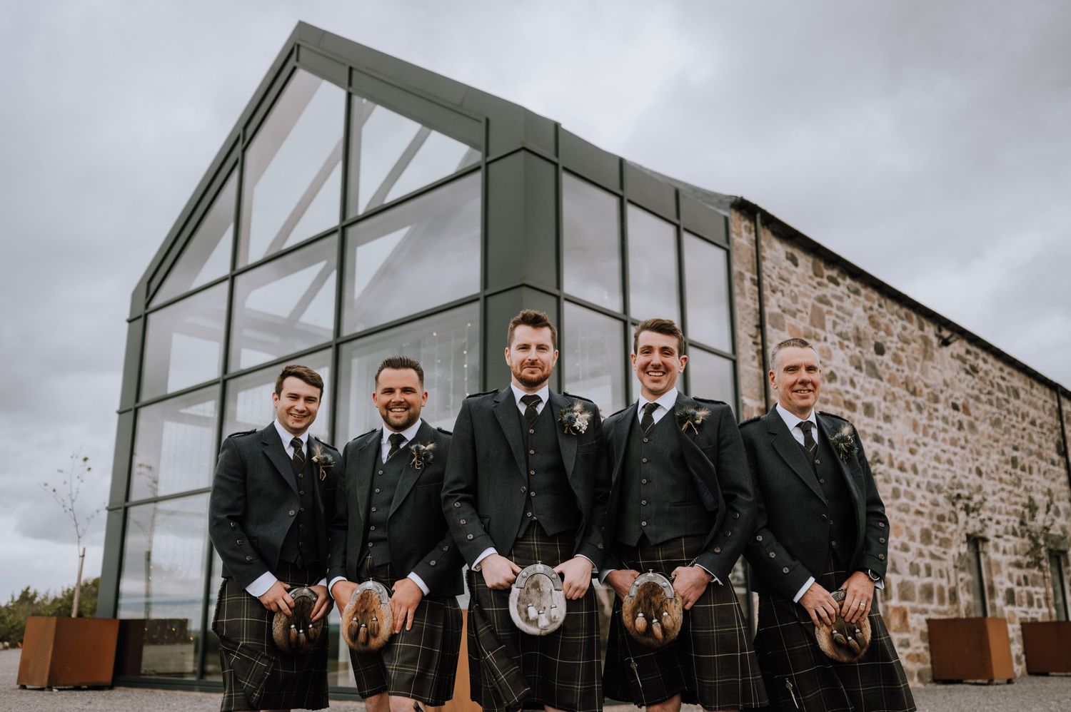 Groomsmen in kilts pose playfully outside modern stone venue with glass architectural features.