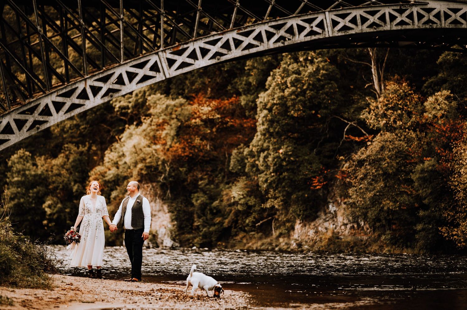 A couple walks hand in hand with their dog beneath a striking iron bridge structure.