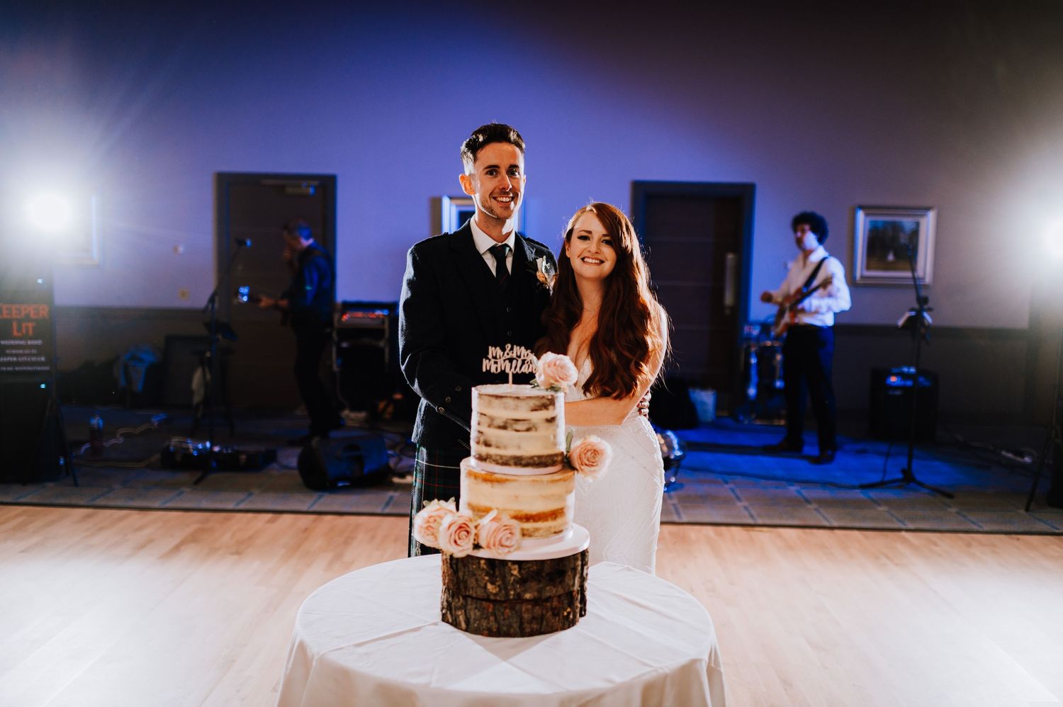 A couple stands with their rustic-style naked wedding cake on a dance floor with a band in background.
