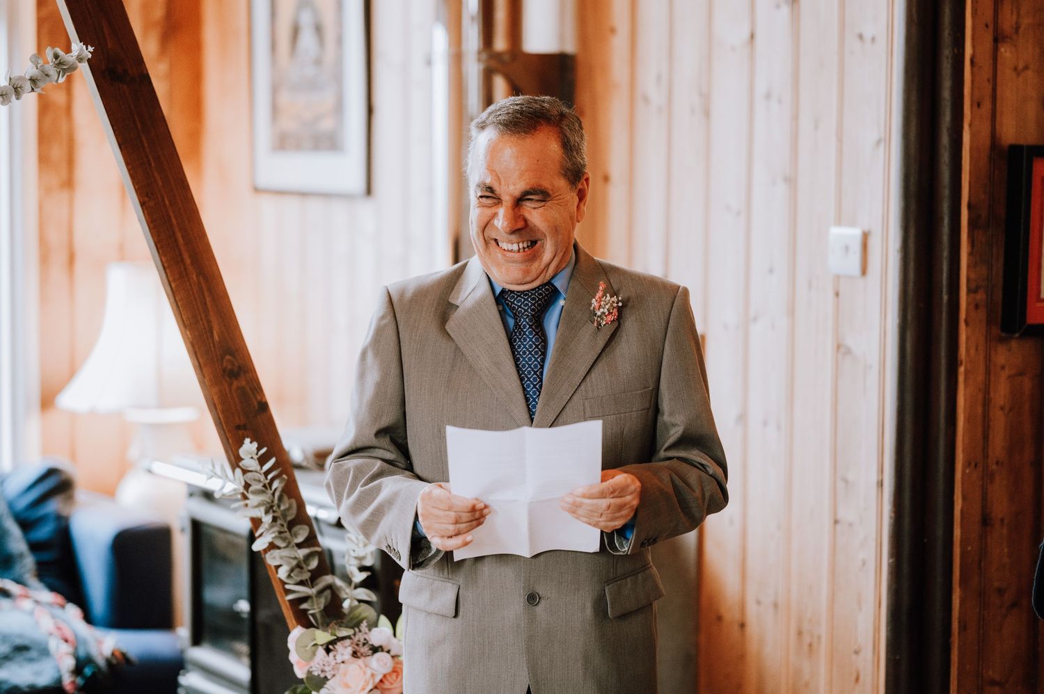A person in a grey suit holds papers while standing in a rustic wood-paneled room with natural light.