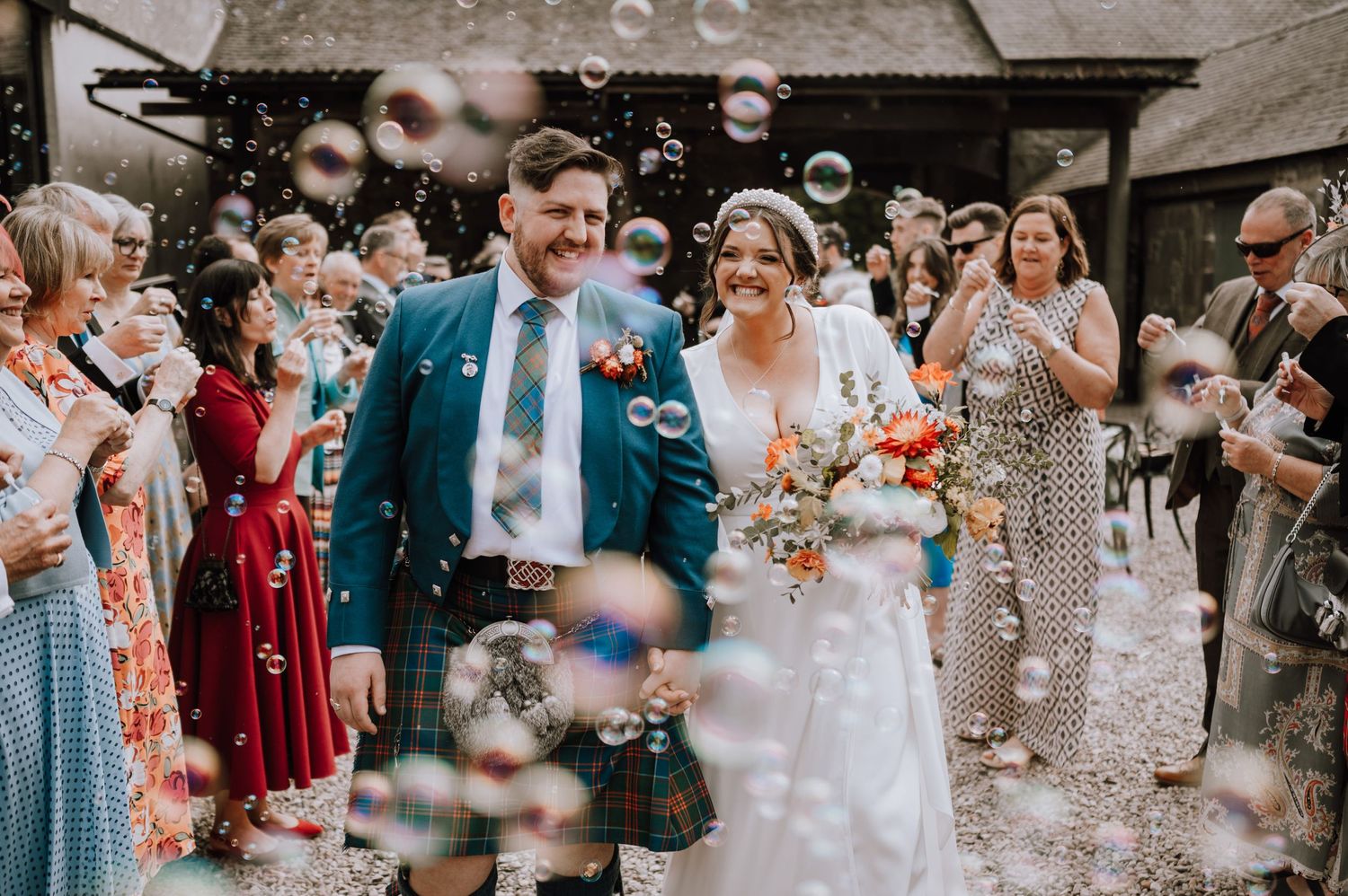 Wedding guests blow bubbles as the couple walks through in Scottish wedding attire.