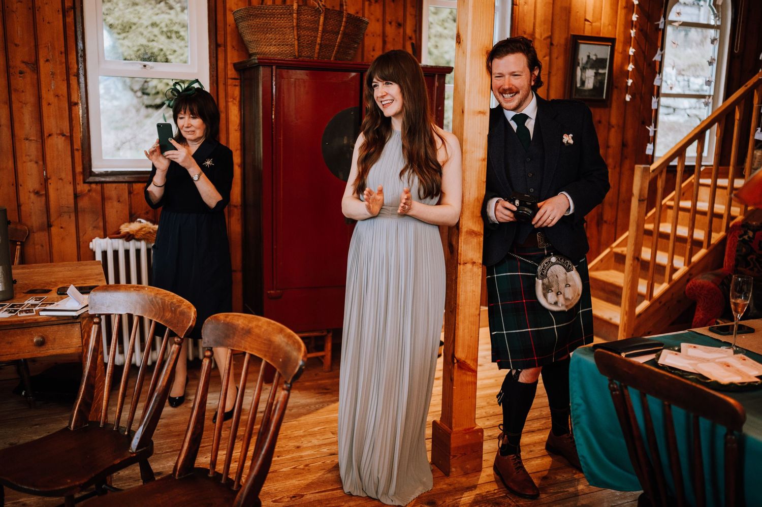 Guests mingle in a rustic wood-paneled room with Scottish Highland wedding attire.