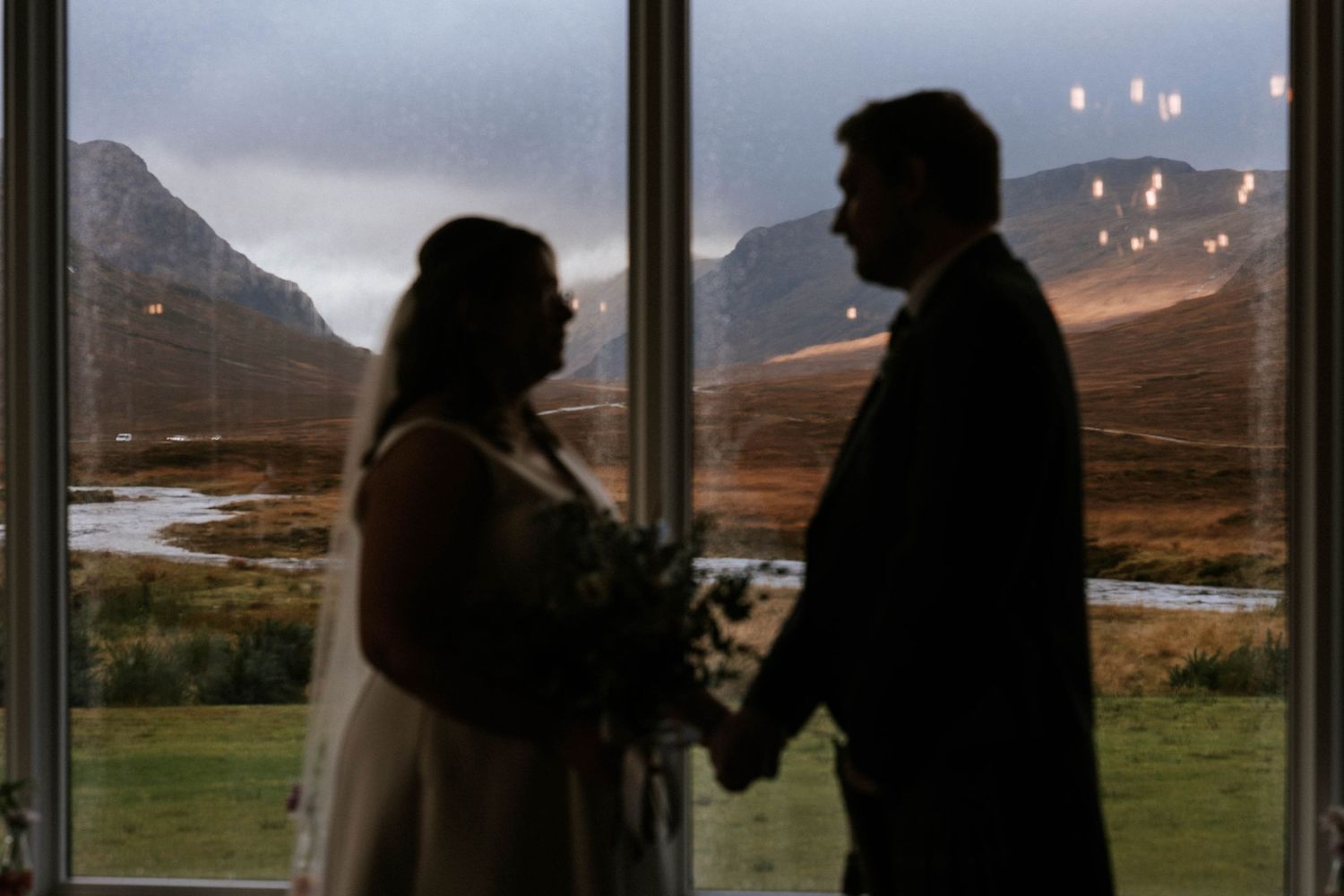 A dark silhouetted wedding party stands in front of large windows overlooking a dramatic mountain landscape at dusk.