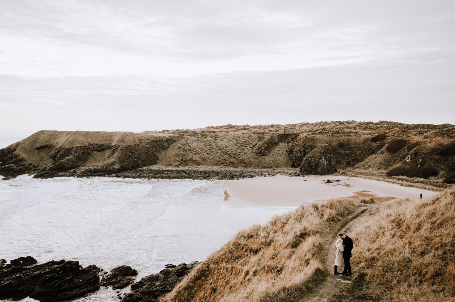 Scenic coastal cliffs and beach view with walking path along rugged shoreline on overcast day.