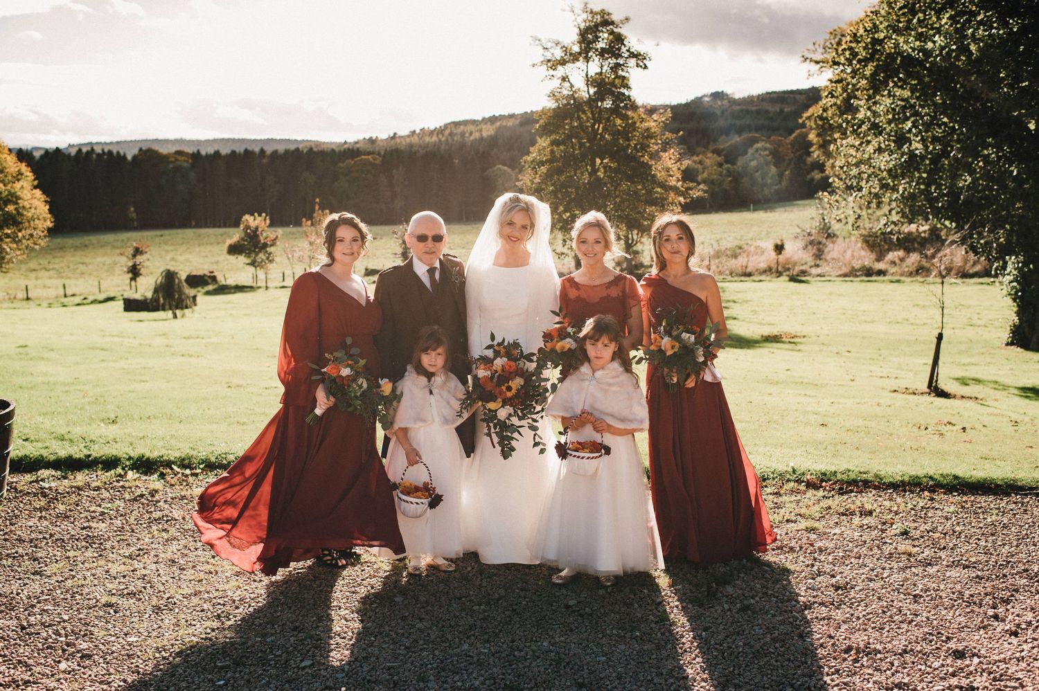 Wedding party poses outdoors with bridal party wearing burgundy dresses against a scenic countryside backdrop.