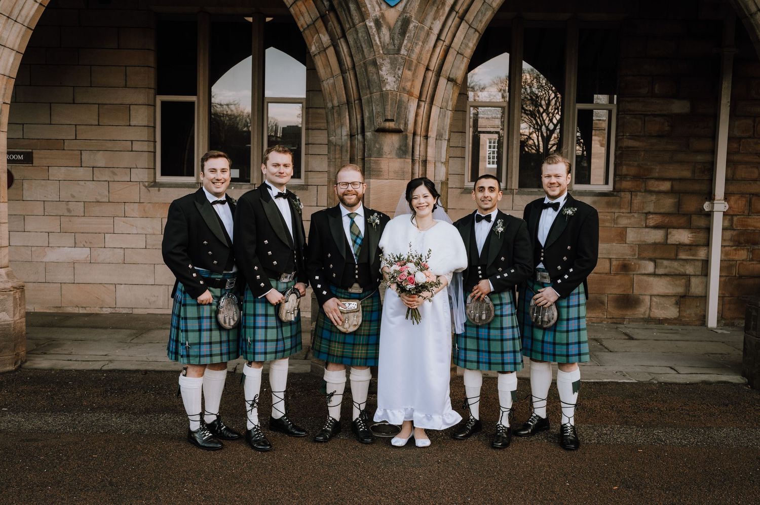 A wedding party in traditional Scottish kilts poses in front of a Gothic stone church entrance.