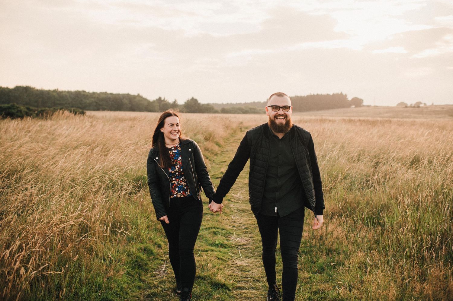 Two people walk hand in hand through a grassy meadow with trees on the horizon.