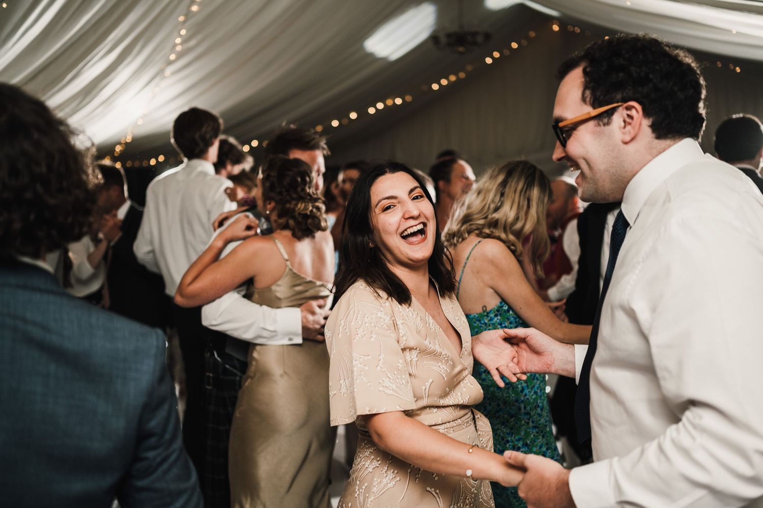 Wedding guests dancing and celebrating at a joyous reception under draped fabric and string lights.