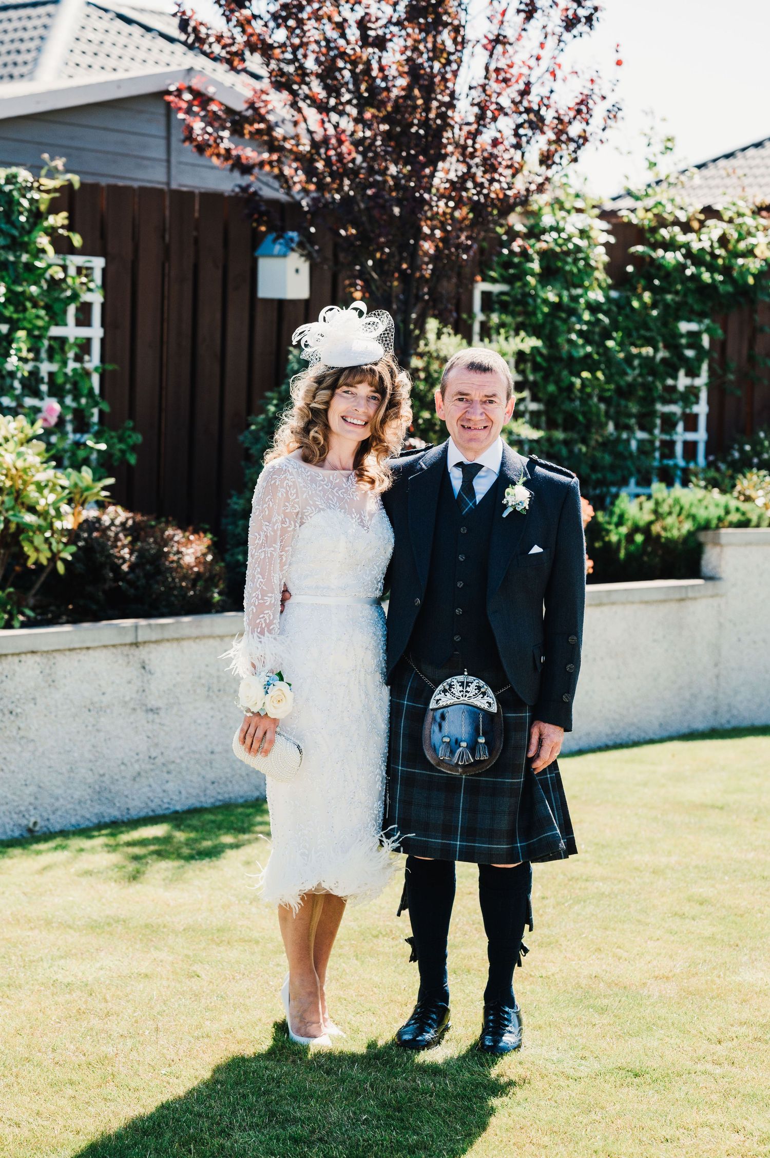 A Scottish wedding couple poses outdoors, with the groom wearing a traditional kilt and the bride in a tea-length lace dress.