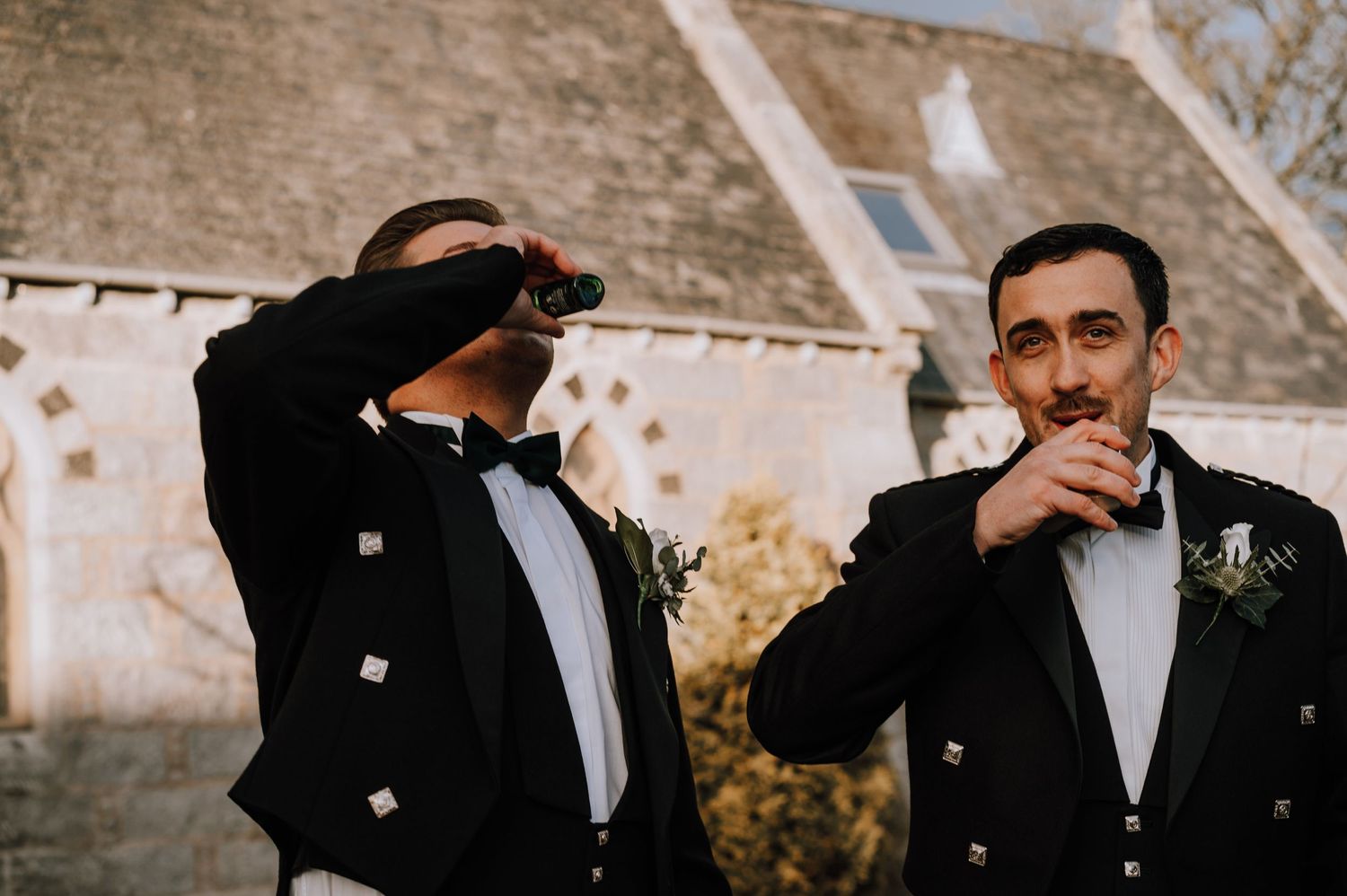 Groomsmen in traditional Scottish attire share a lighthearted moment outside.