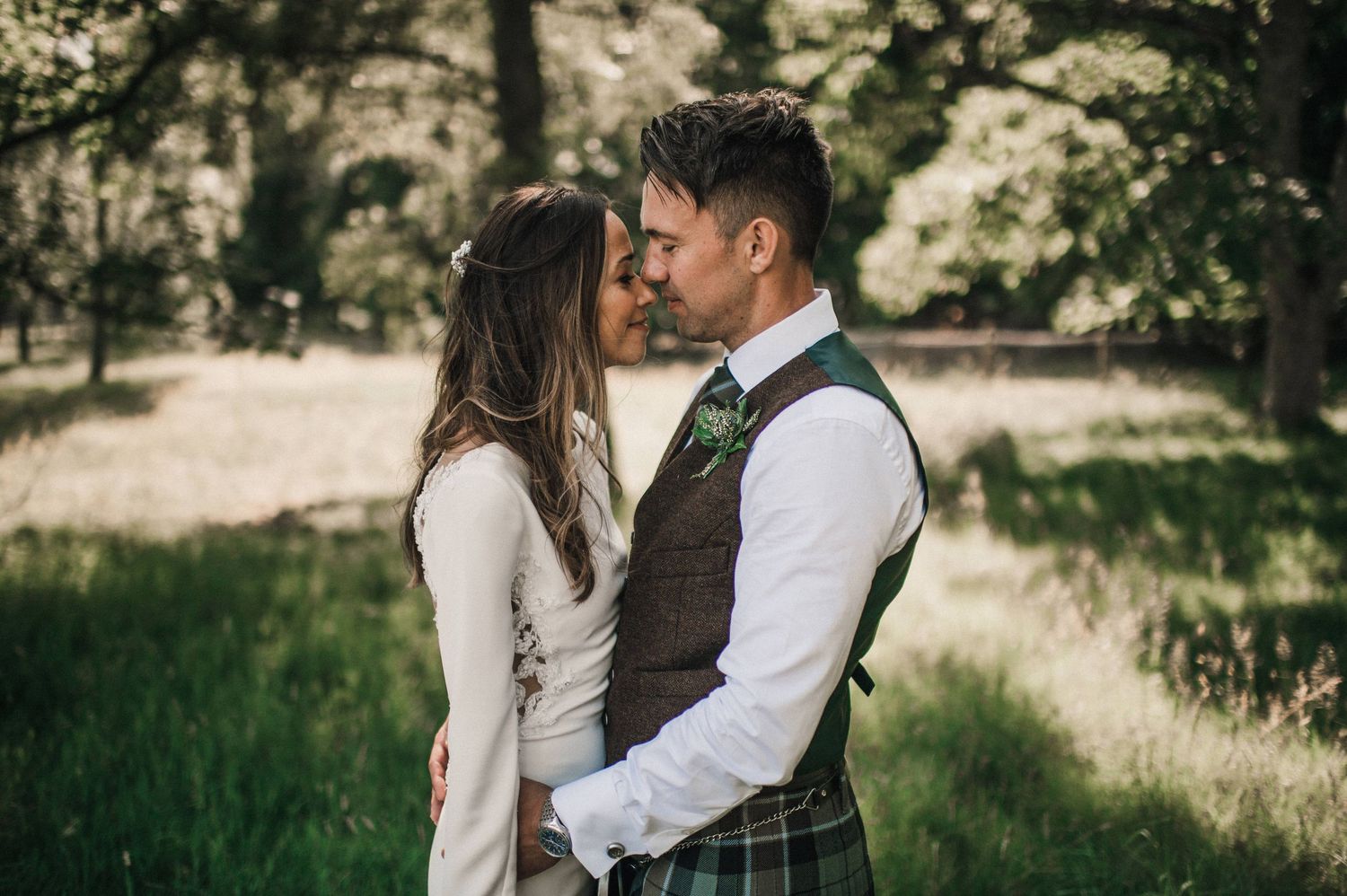 Intimate moment between newlyweds in Scottish wedding attire against sunlit forest backdrop.