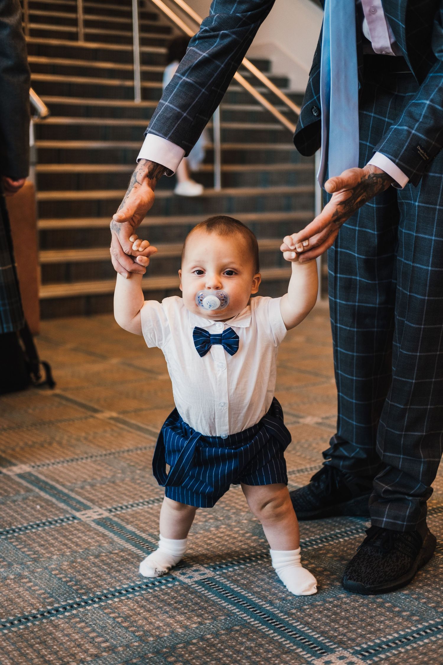 Baby in white shirt and bow tie holds hands with adults while learning to walk.