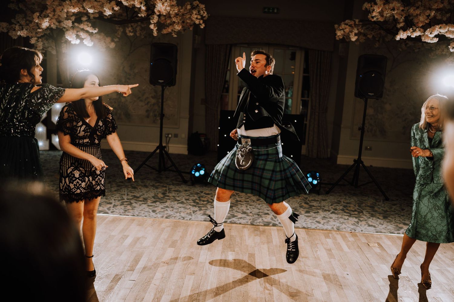 A person in a kilt performs an energetic dance move at a wedding reception.