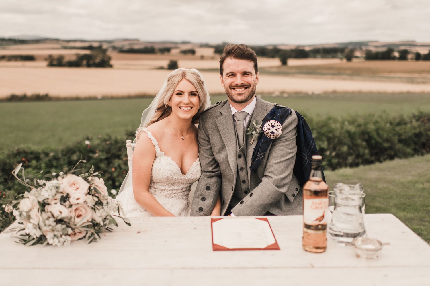 A couple poses behind a white signing table at their outdoor wedding venue.