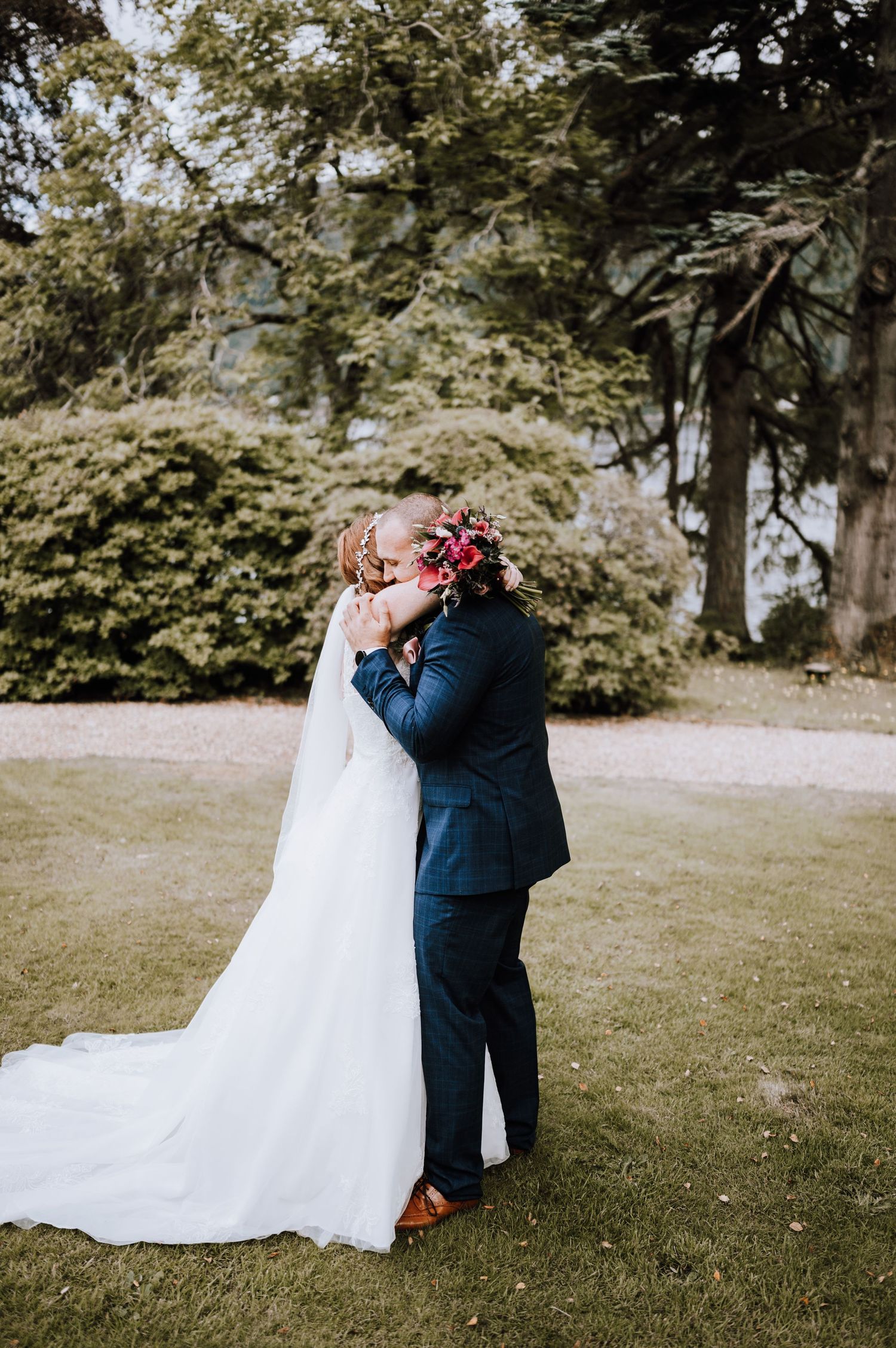 Newlyweds embrace on lawn with trees and water in background.