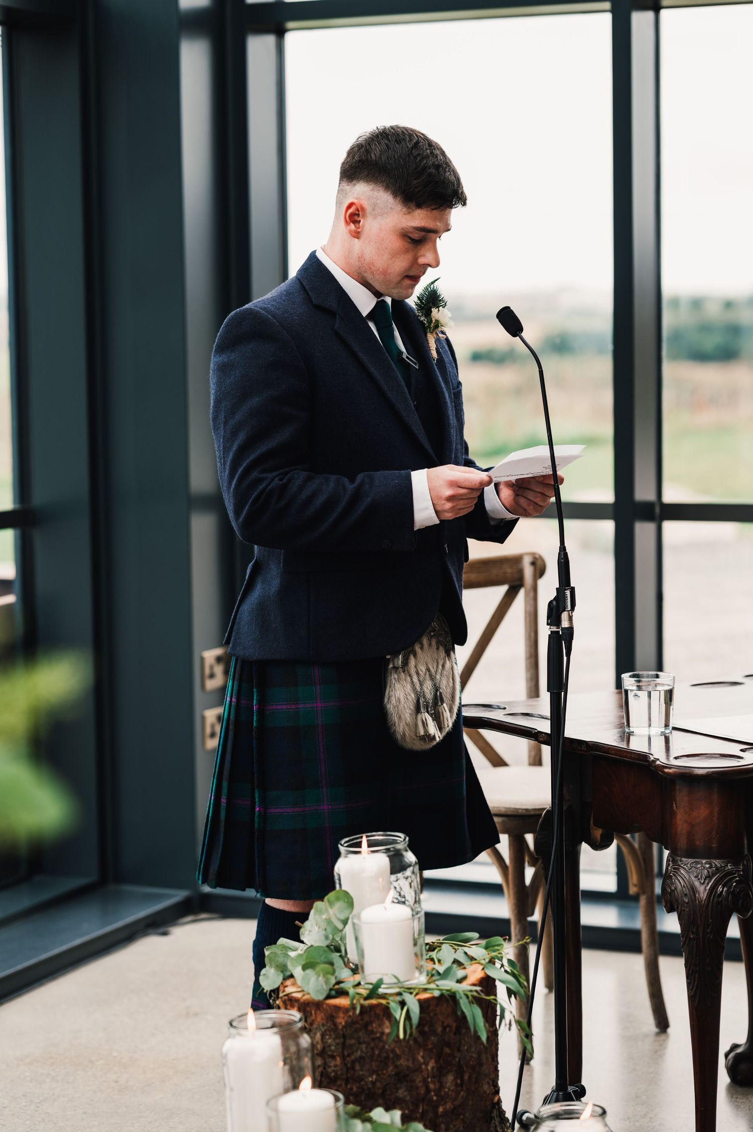 Speaker in traditional Scottish formal wear giving a speech at wedding reception.