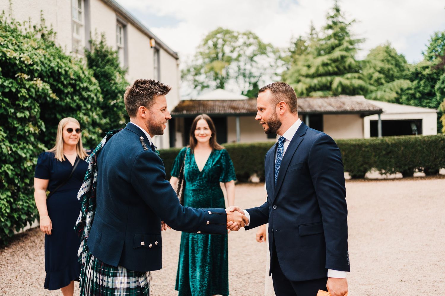 Formal handshake between wedding attendees wearing kilts and suits on gravel path.