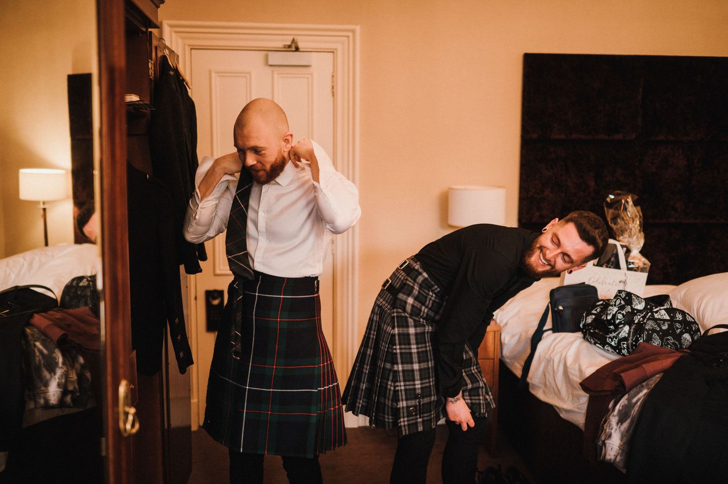 Two men in kilts prepare for a formal Scottish event in a cozy bedroom setting.