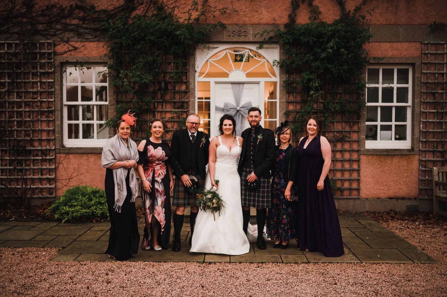 Wedding party members gather for a formal photo in front of an ivy-covered brick building.