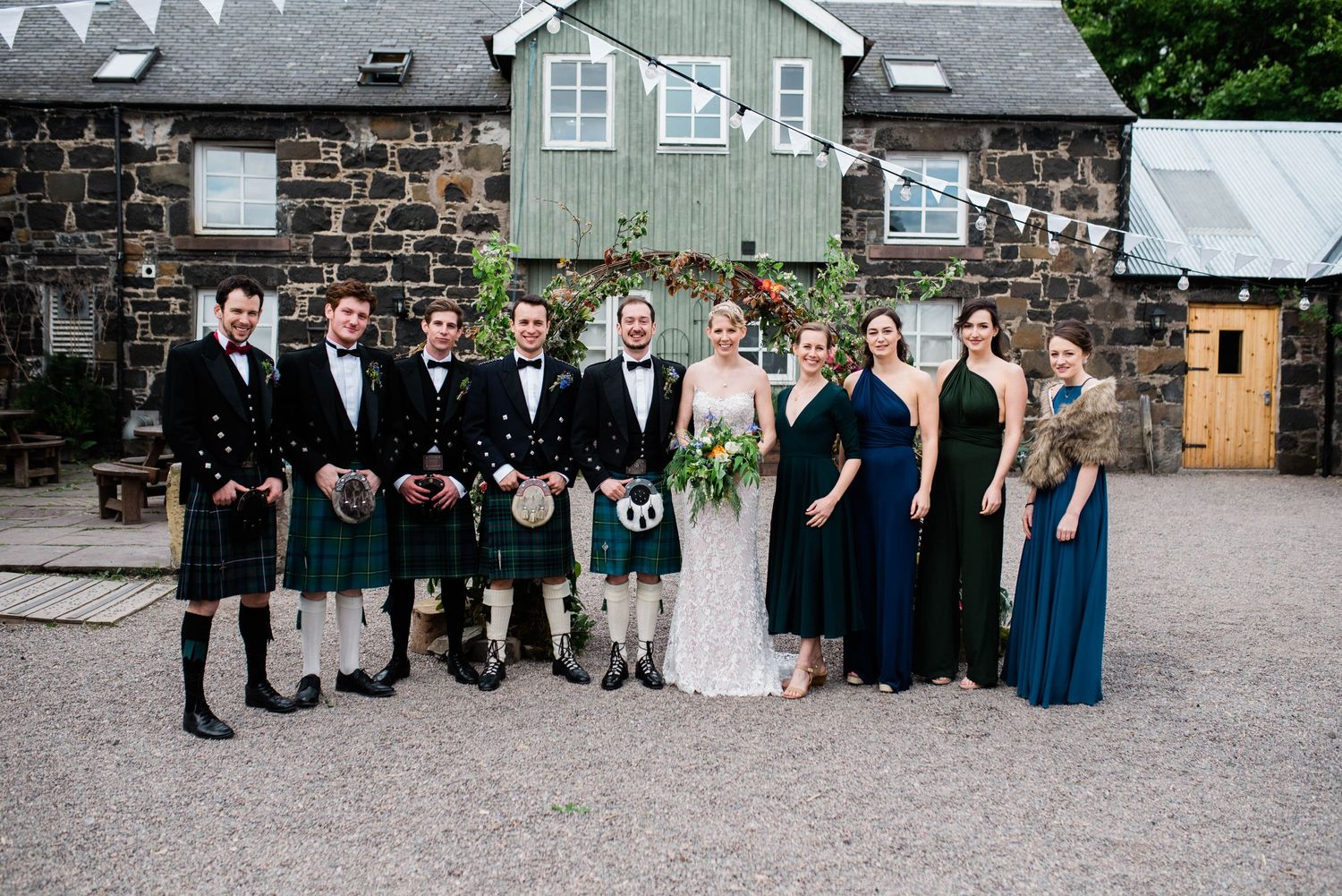 Large wedding party in Scottish formal wear and evening gowns gathered outside a stone and sage green building.
