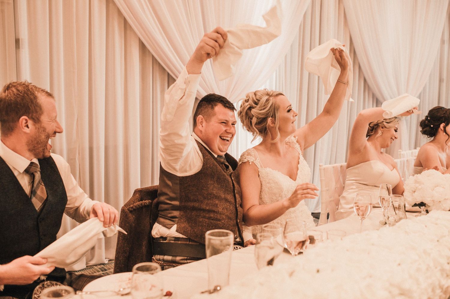 Wedding party celebrates with raised arms at elegant white-decorated reception venue.