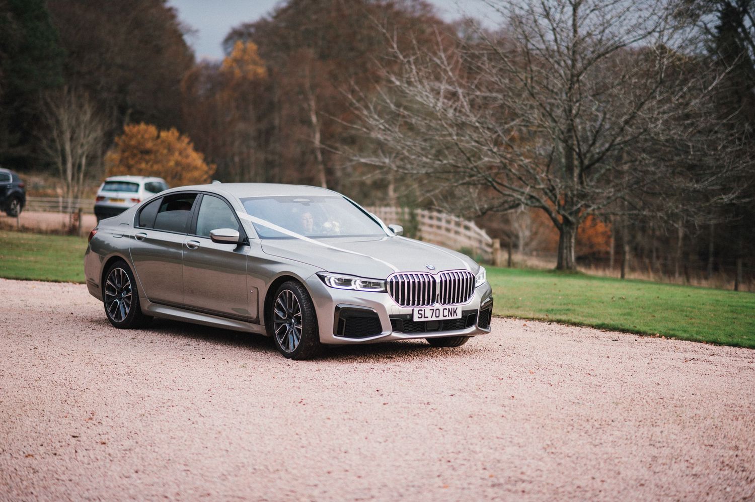 A silver BMW 7-series luxury car parked on a gravel driveway with autumn trees in background.