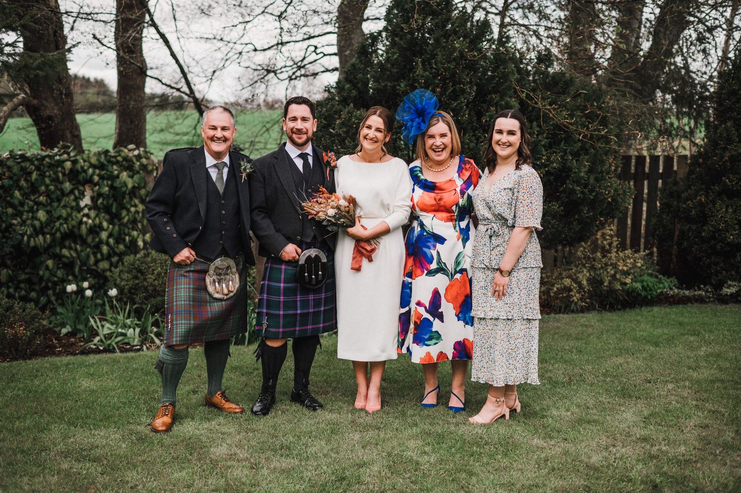 Group photo of wedding party members in mix of kilts and formal attire standing in garden setting.