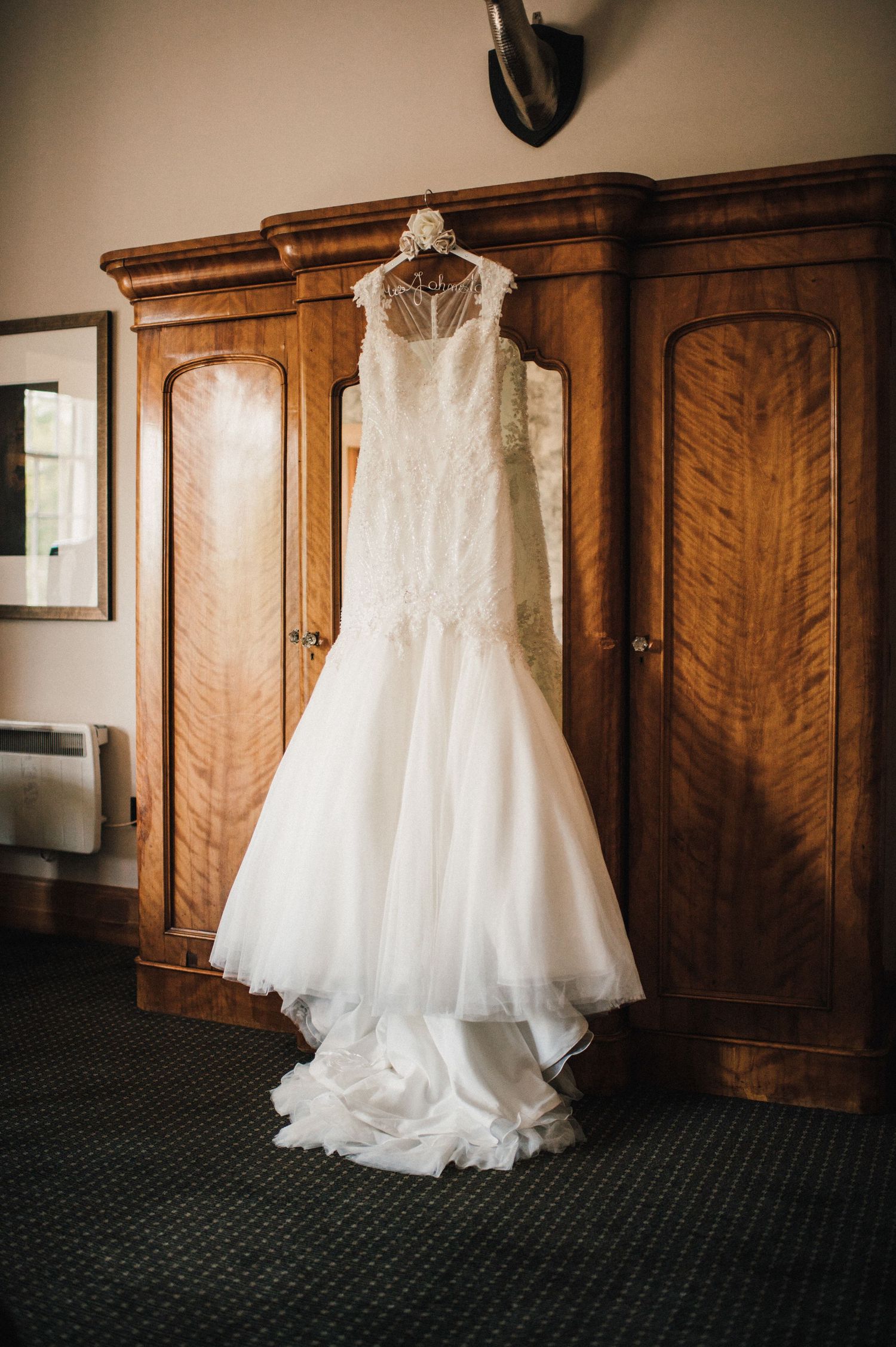 An elegant white wedding gown displayed on a hanger against a wooden wardrobe.