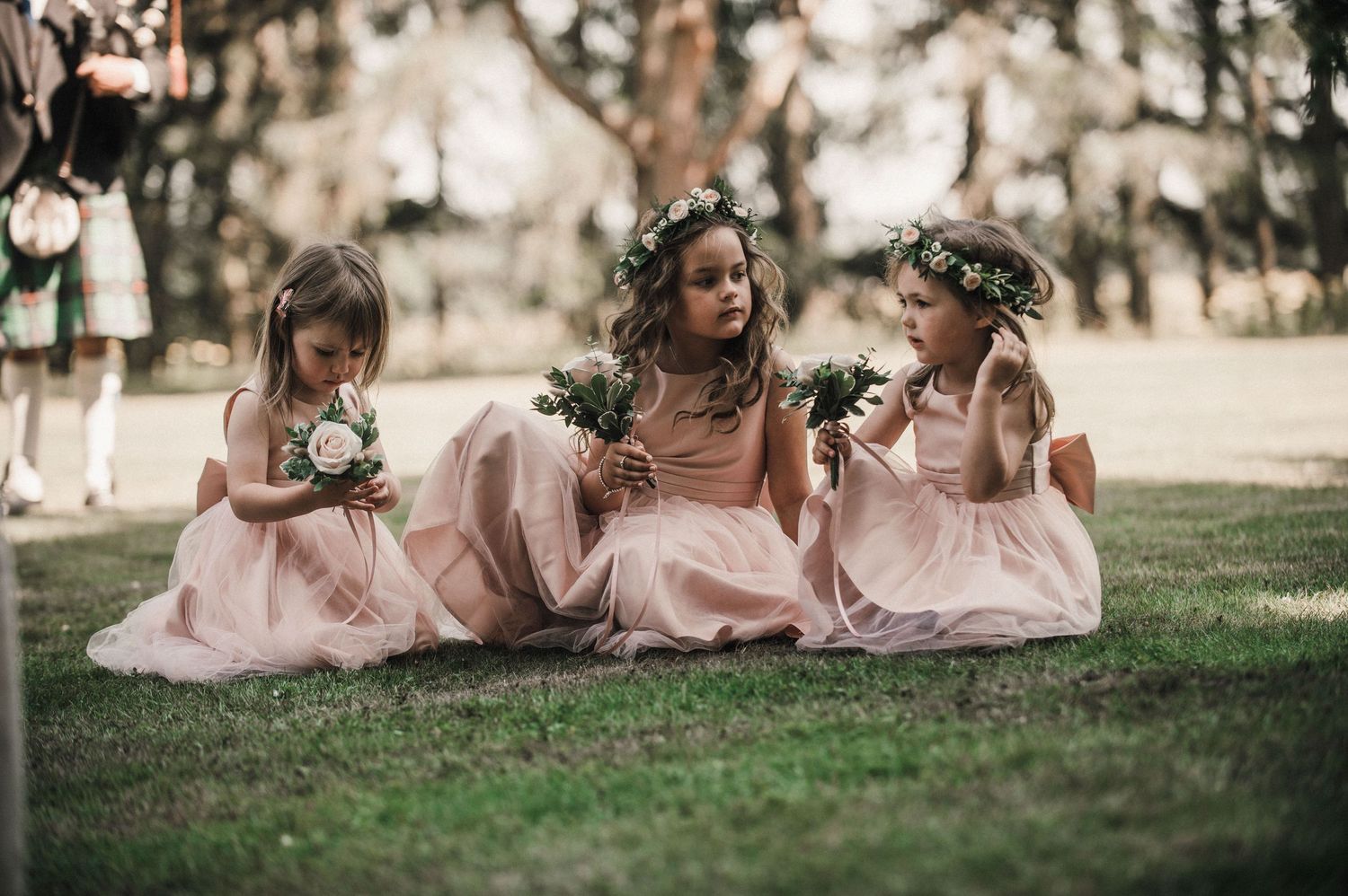 Three young attendants in pink dresses sit on grass holding flowers.