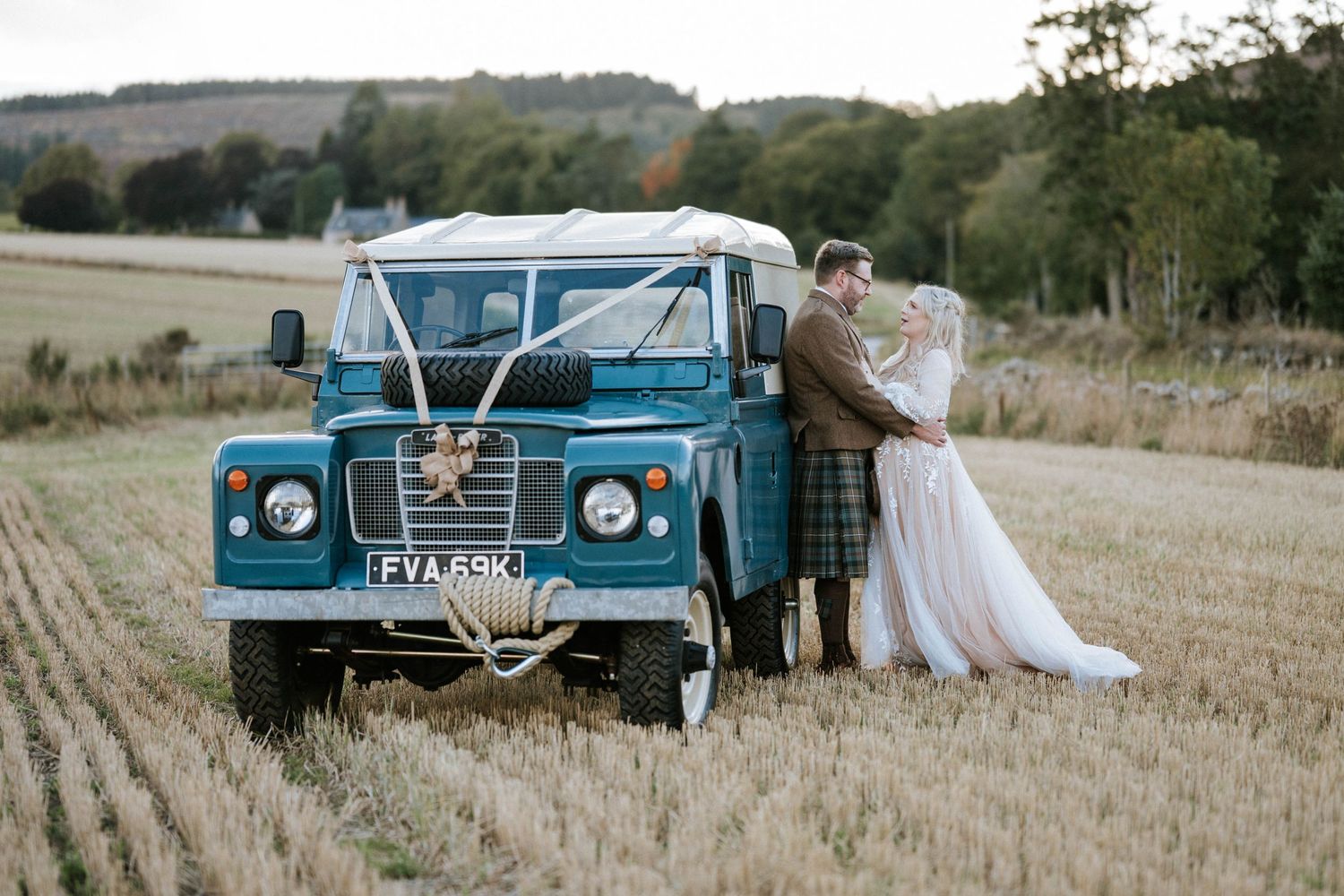 Vintage blue Land Rover decorated with wedding ribbons parked in a harvested field.