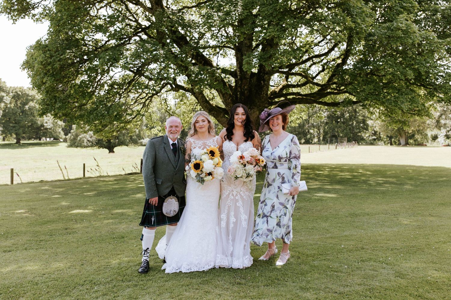 Wedding party and guests gather for outdoor group photos under a sprawling tree.