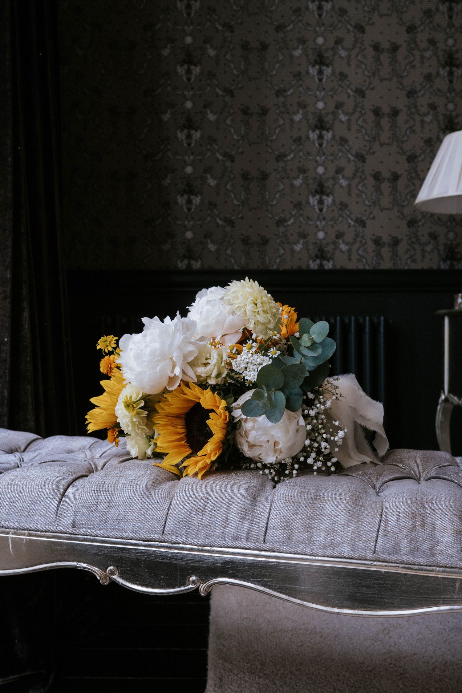 Wedding bouquet with sunflowers and white peonies resting on gray upholstered furniture.
