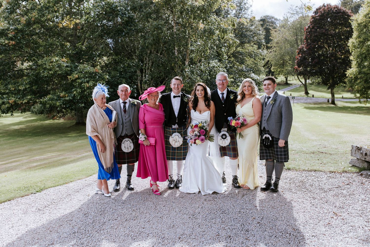 Wedding party members in colorful formal attire and kilts pose together on garden grounds.
