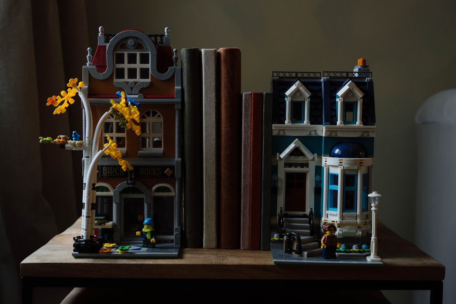 LEGO modular buildings used as bookends with books between them on a wooden shelf in dim lighting.