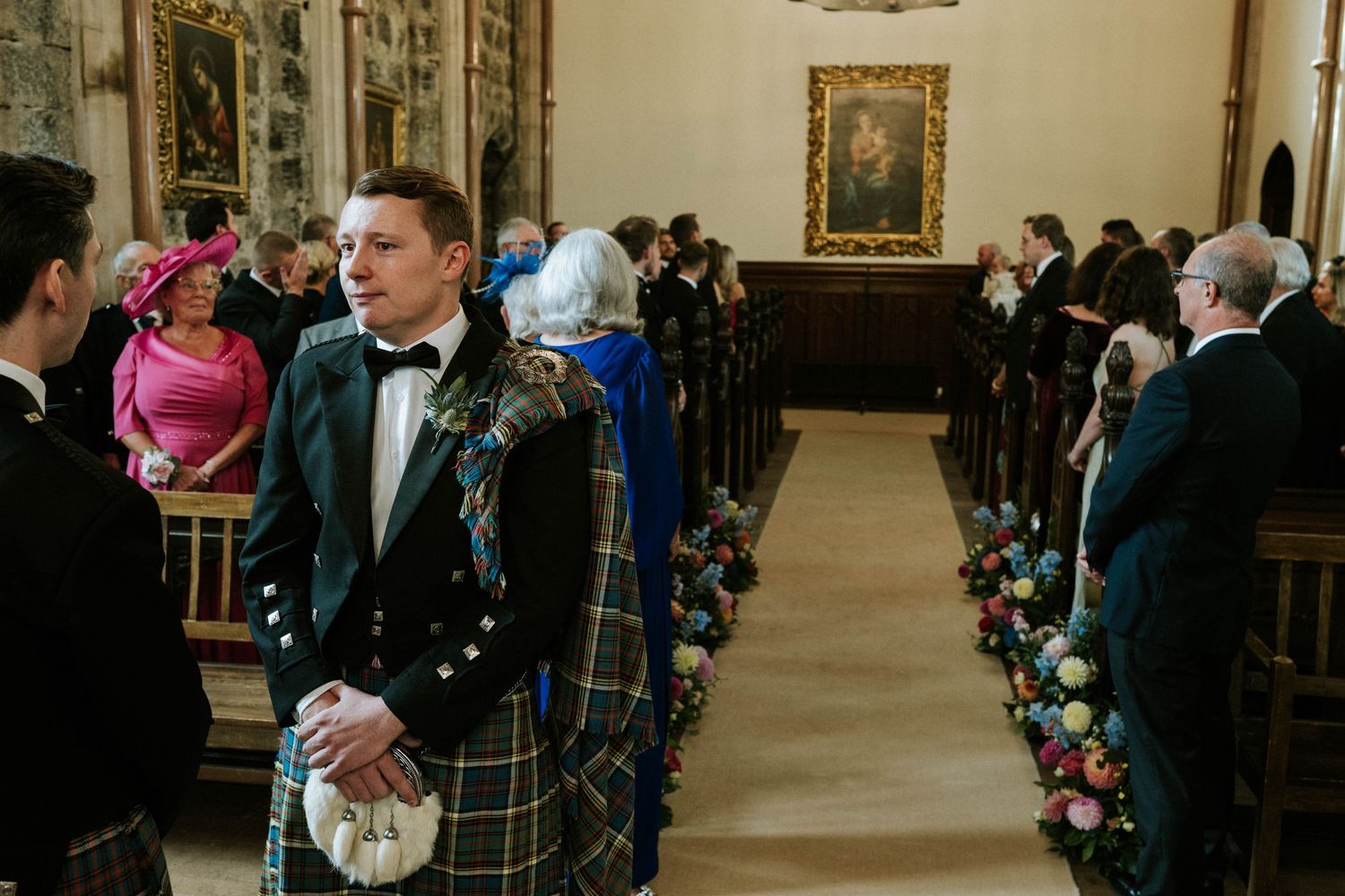 Wedding ceremony guests gather in a grand hall with traditional Scottish attire and decorative floral arrangements.