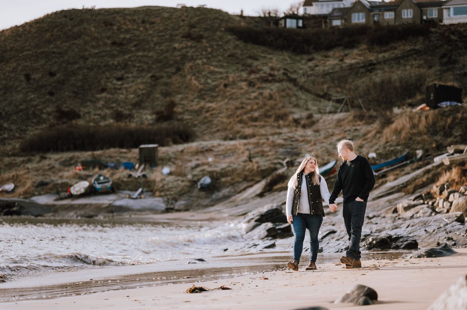 A panoramic view of people walking along a rocky coastal beach with white houses visible on the hillside.