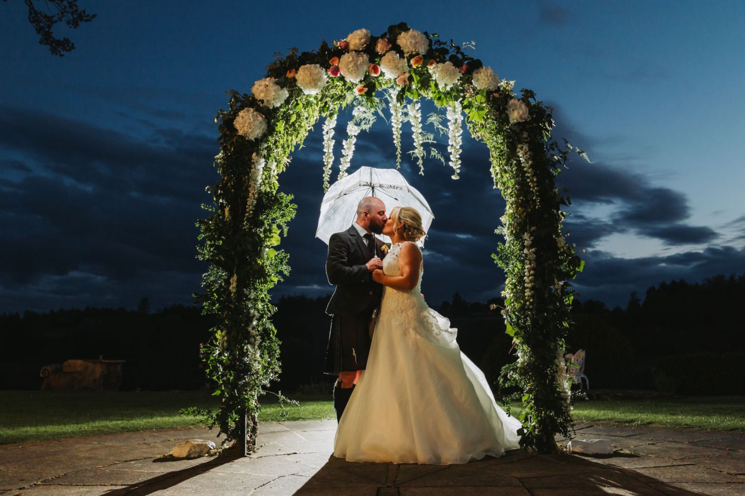 A romantic wedding couple embraces under an illuminated floral arch at dusk with a dramatic evening sky backdrop.