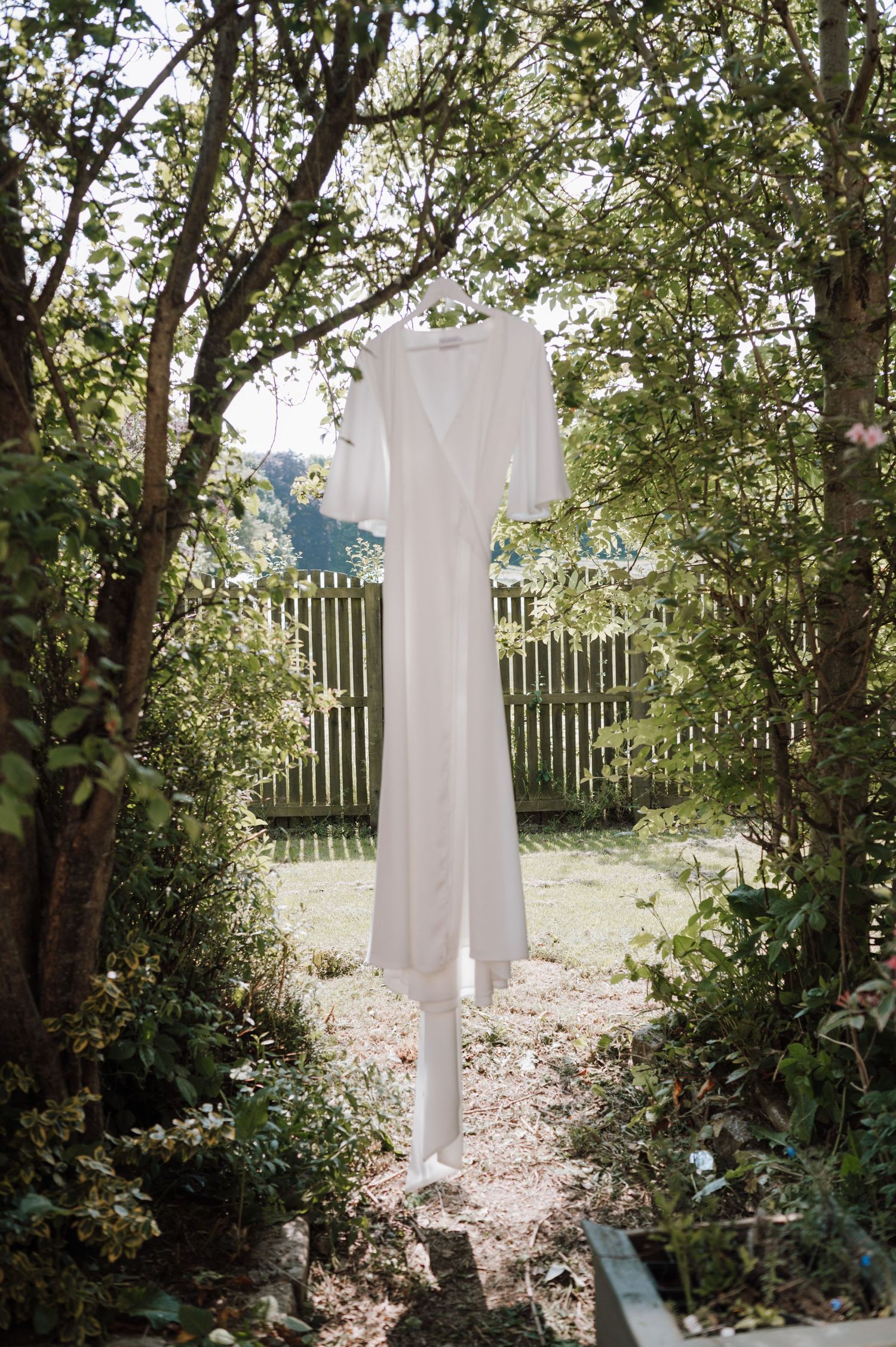 A white wedding dress hanging outdoors between trees with a wooden fence in the background.