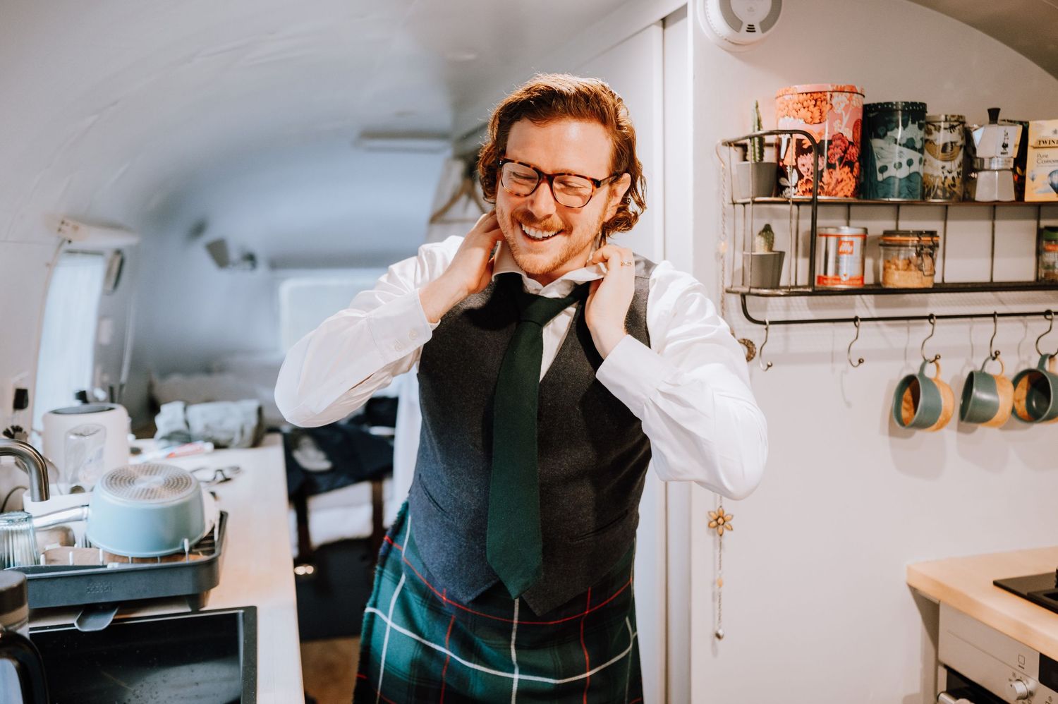 Person in formal Scottish attire adjusting outfit while smiling in small room.