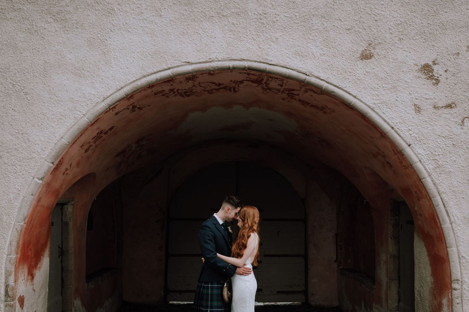 Wedding couple embraces under stone archway during intimate moment.