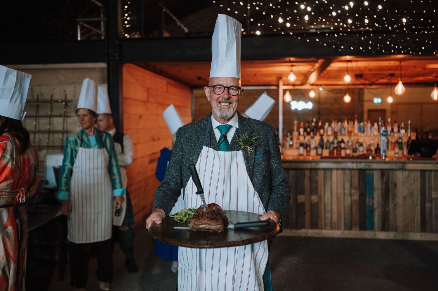 A server in an apron and glasses presents a dish at a rustic wedding reception venue.