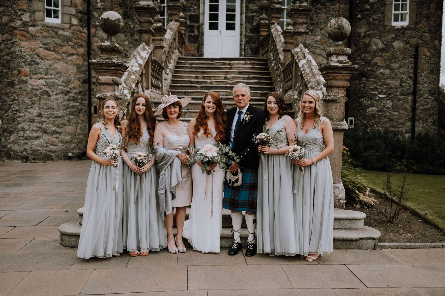 A wedding party in traditional Scottish attire including kilts and light gray bridesmaid dresses poses on stone steps.