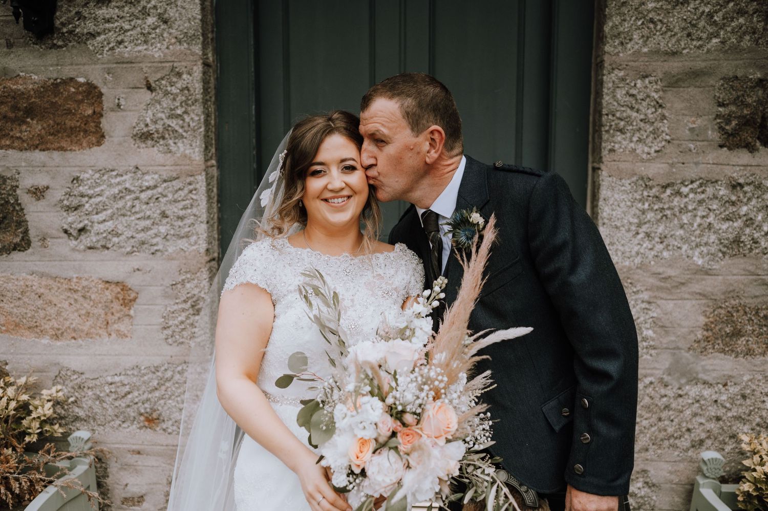 A tender moment between newlyweds holding a romantic dried flower bouquet against a stone wall.