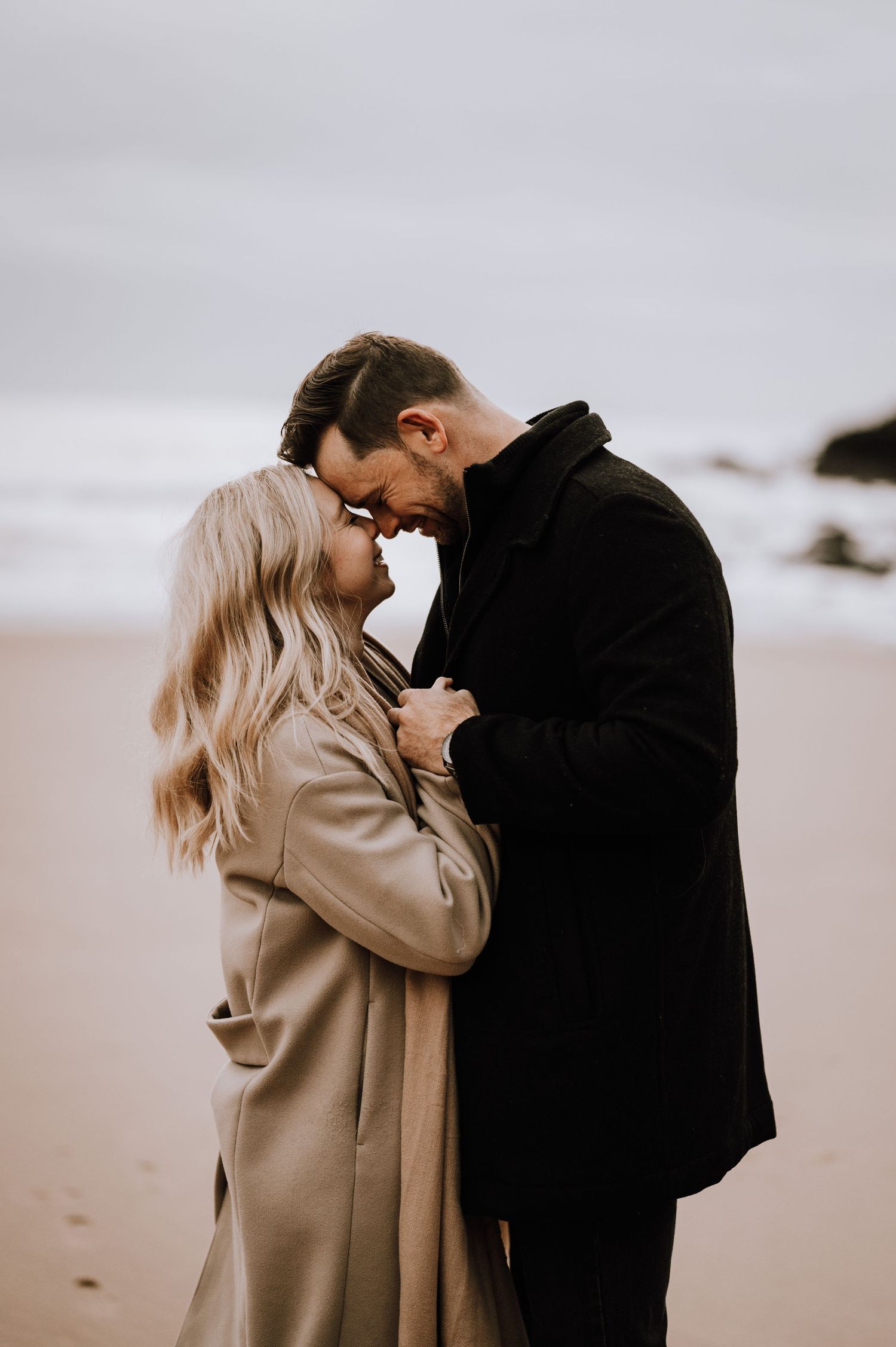 Romantic couple in winter coats sharing an intimate moment on a cloudy beach.
