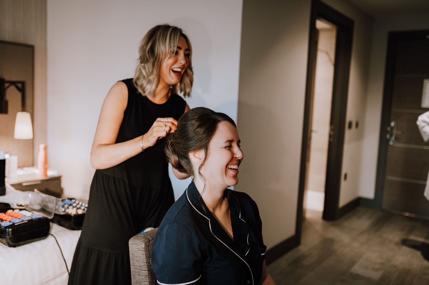 A stylist works on styling hair during wedding preparation in a dimly lit room.