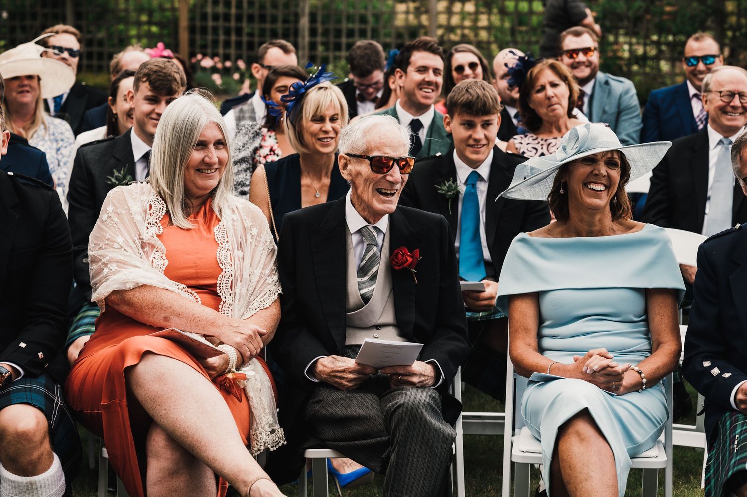 Wedding guests in formal attire seated on white chairs during outdoor ceremony.