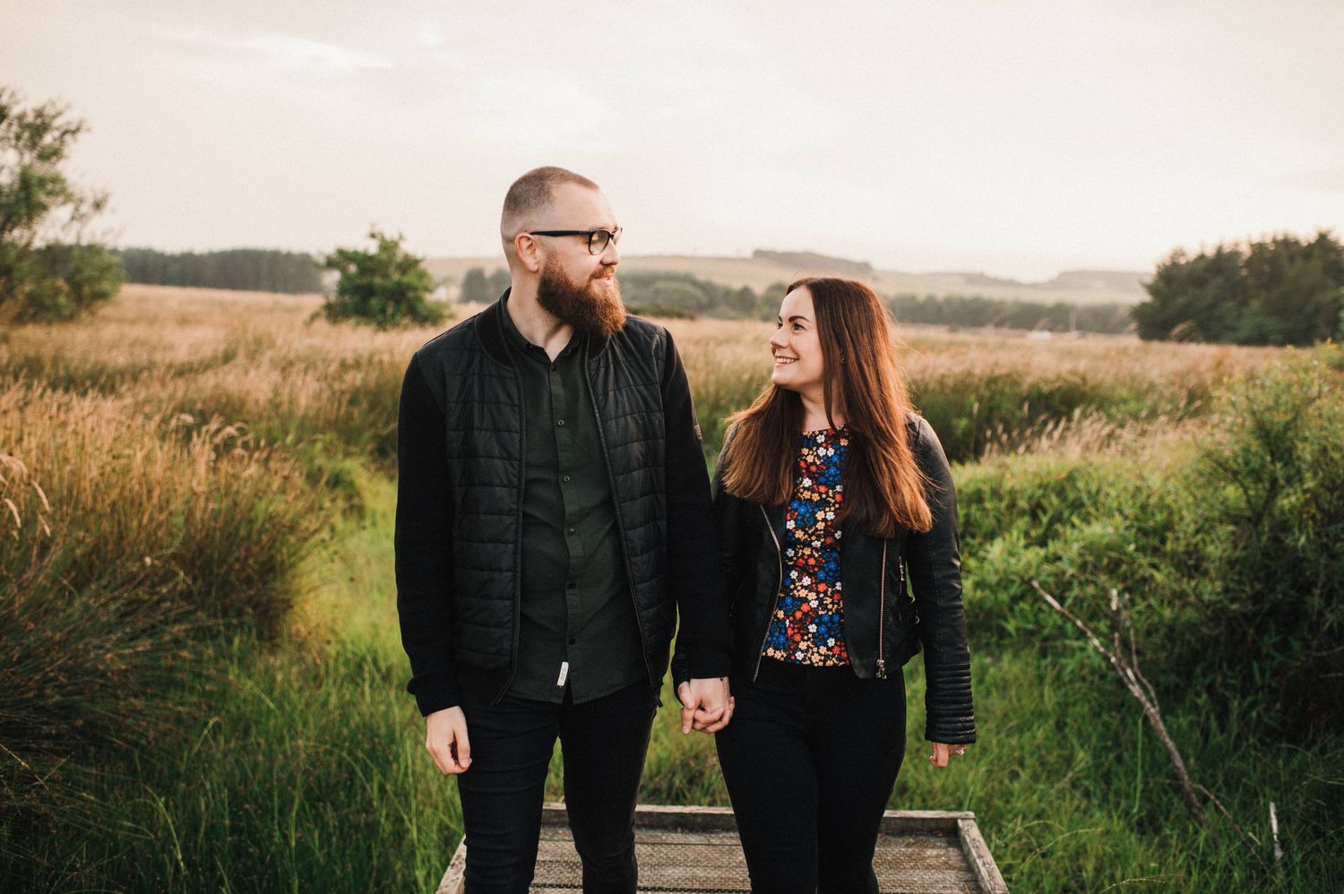 Couple walking along wooden boardwalk through natural grassland setting.