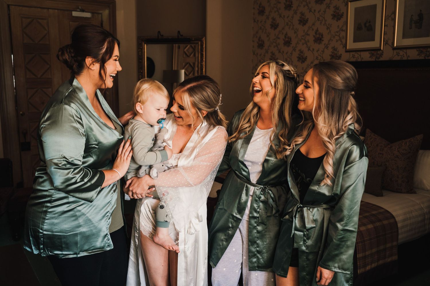 Candid moment of wedding party in sage green robes sharing laughter in bedroom.