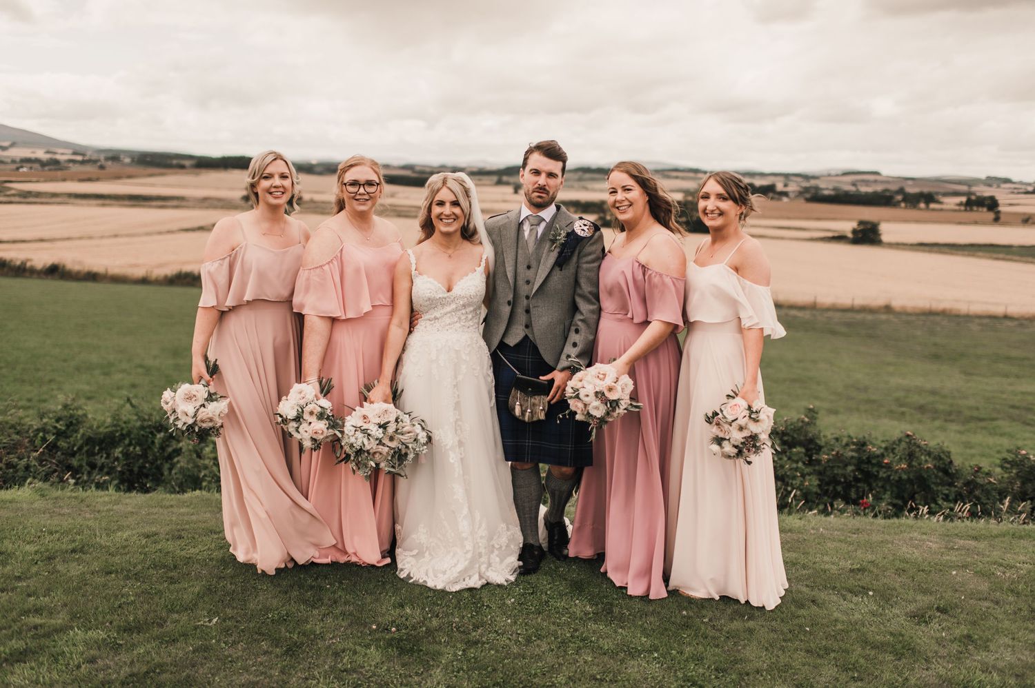 Wedding party with bridesmaids in flowing pink dresses and groom in traditional kilt pose in countryside setting.
