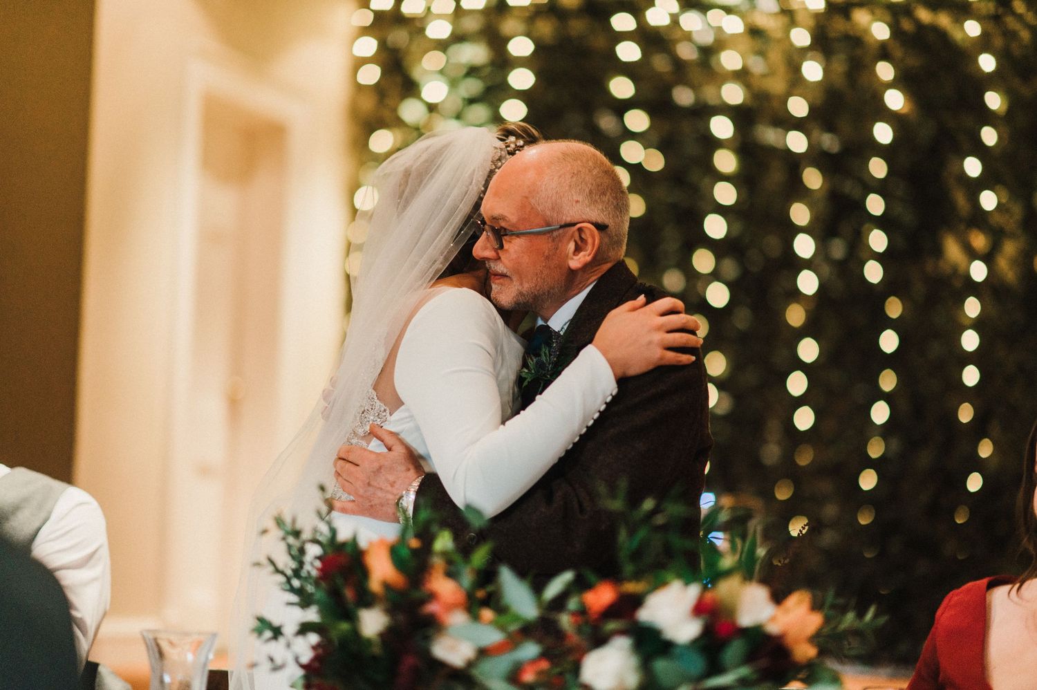 An emotional wedding reception moment captured as two people embrace under warm twinkle lights and floral decorations.